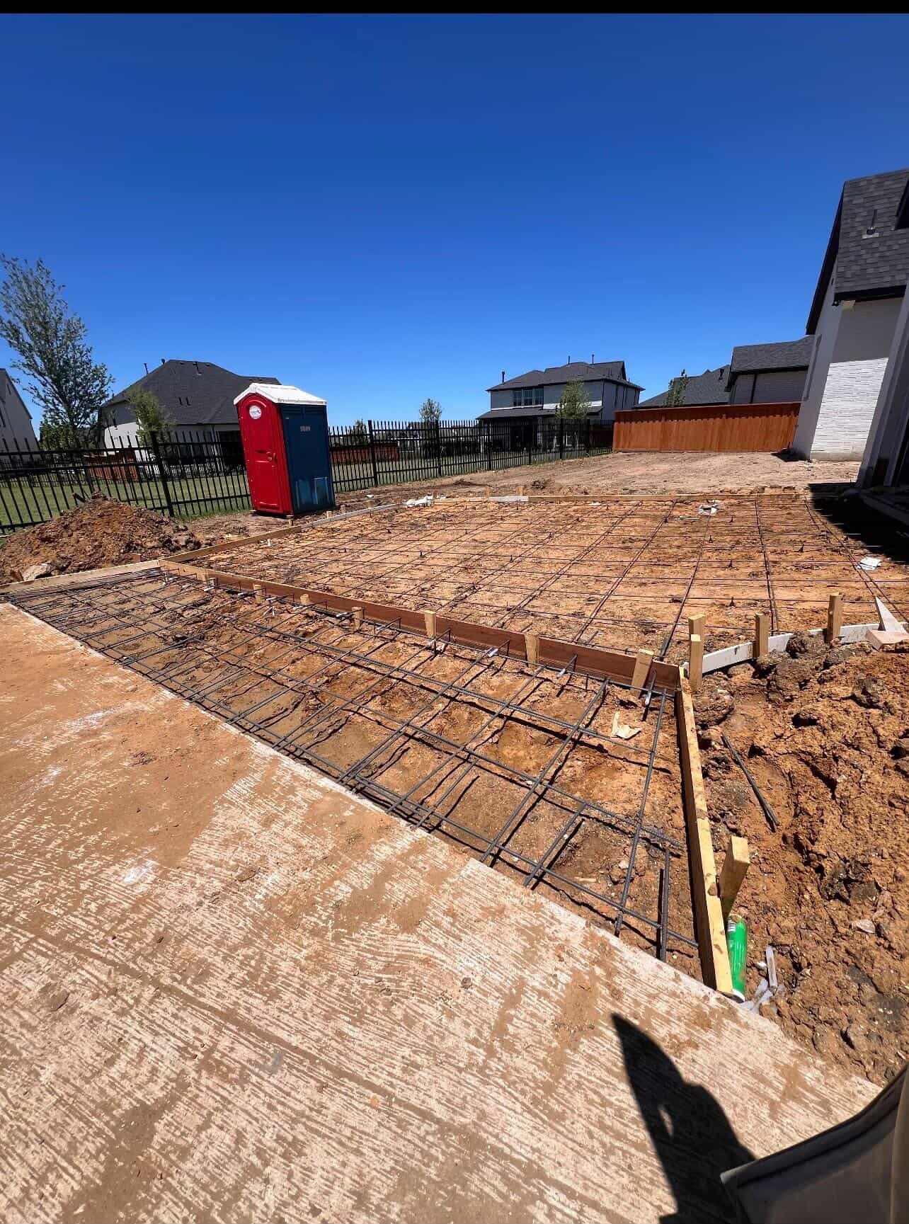 Construction site with steel reinforcement bars laid on dirt foundation, residential houses in background under clear blue sky