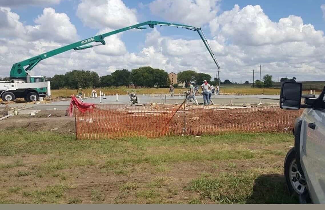 Concrete pump truck spraying foundation at construction site with safety fencing and workers present