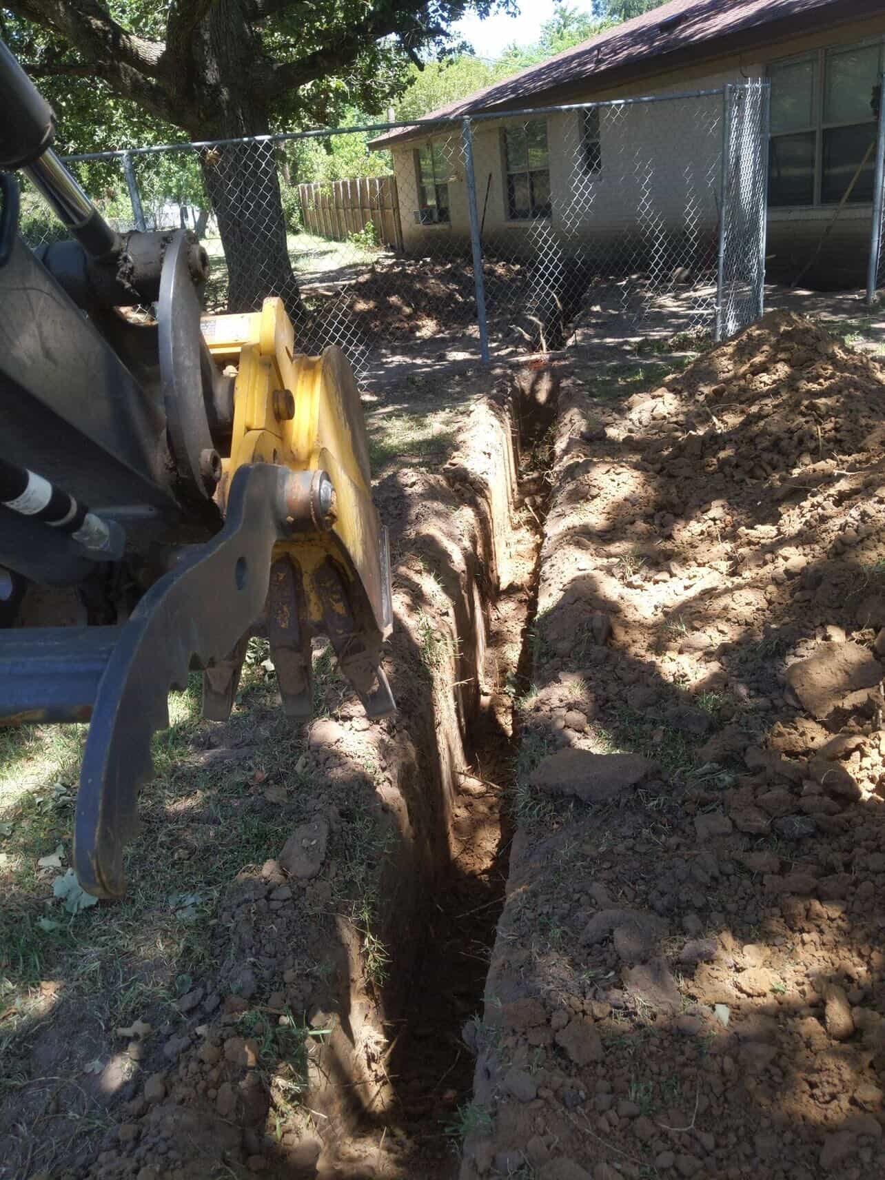 Yellow skid-steer loader digging a trench next to a residential house with metal fencing and large tree