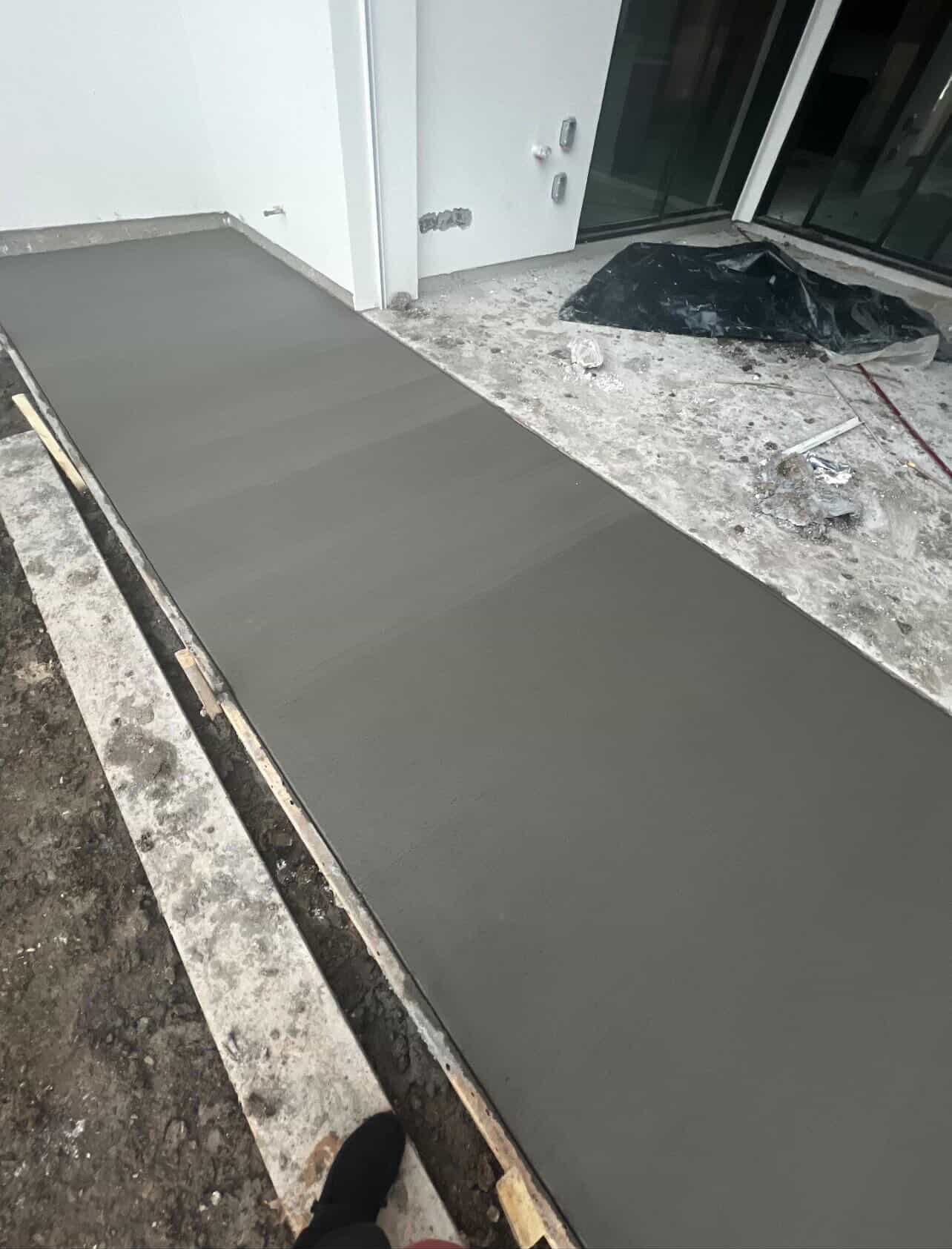 Fresh gray concrete being poured and smoothed on a residential exterior surface near white walls and doors