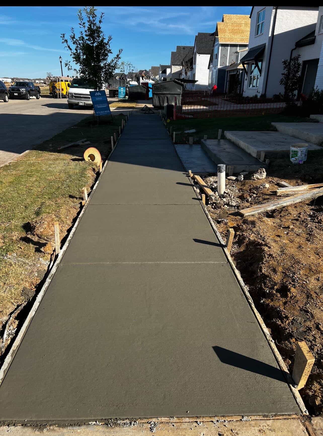 Freshly poured concrete walkway with wooden forms along residential street, homes and construction equipment visible in background