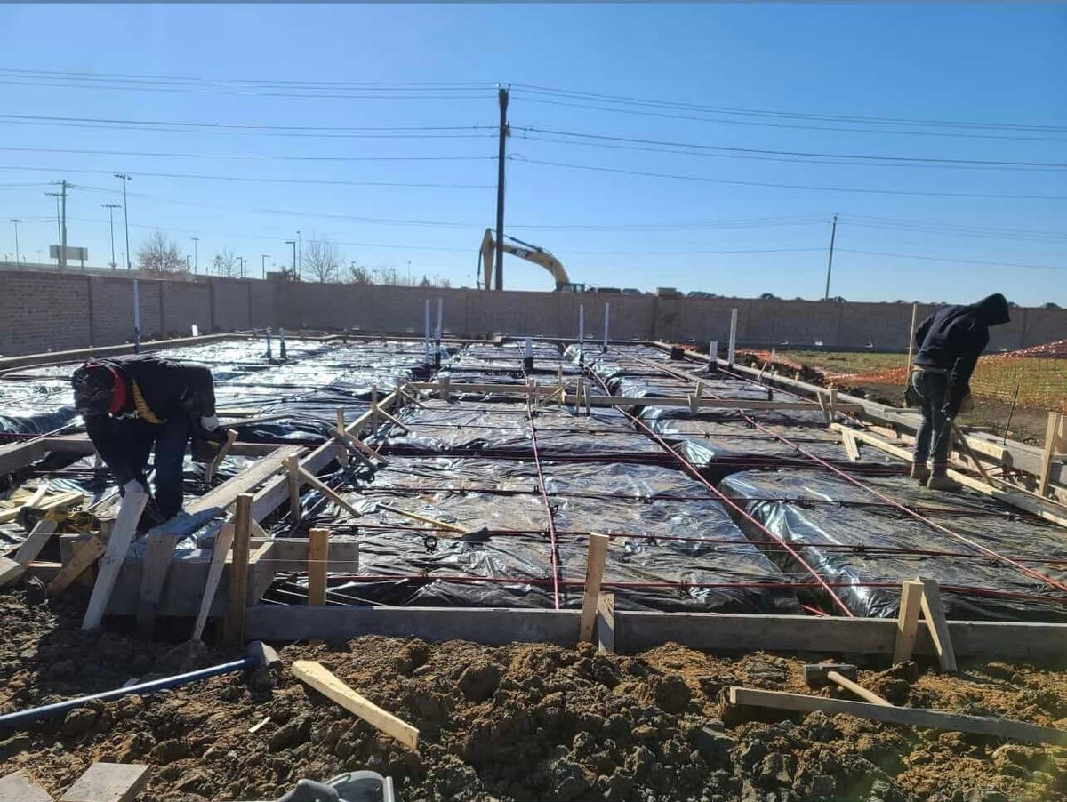 Construction site with concrete foundation, wooden framing, metal poles, and workers on cleared land under blue sky