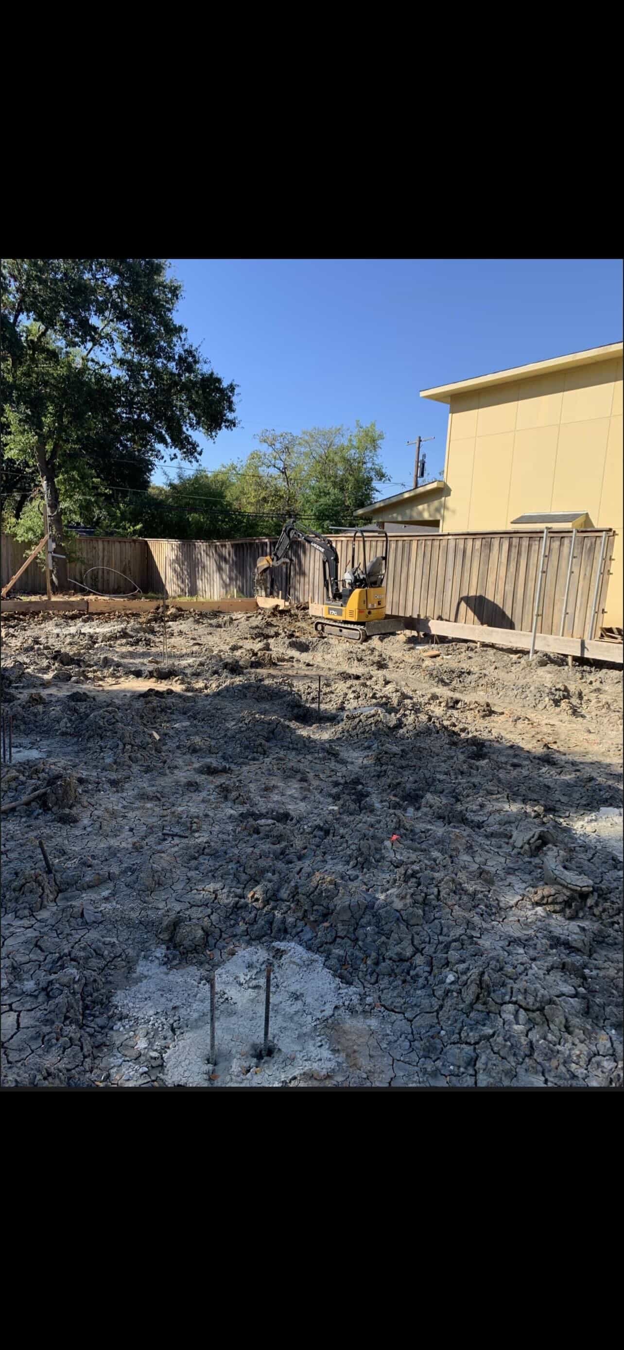 Backyard construction site with excavator, metal fencing, yellow house, and bare dirt ground on sunny day